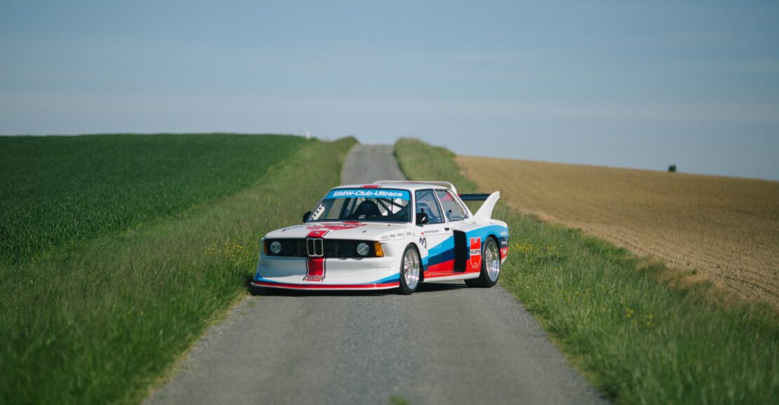 BMW E21 Groupe 5 réplique aux couleurs blanches, bleues et rouges sur une route de campagne